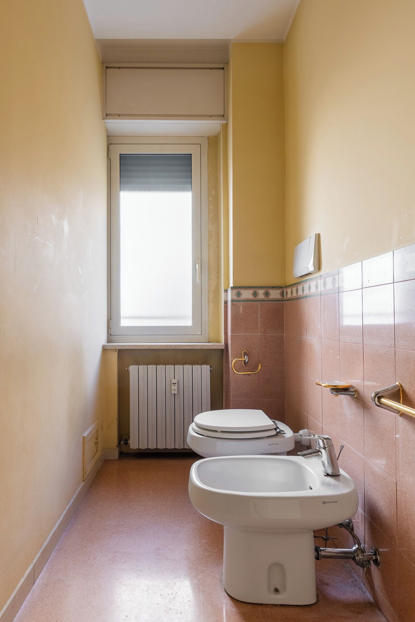 A narrow bathroom with two toilets, a sink, and a window.