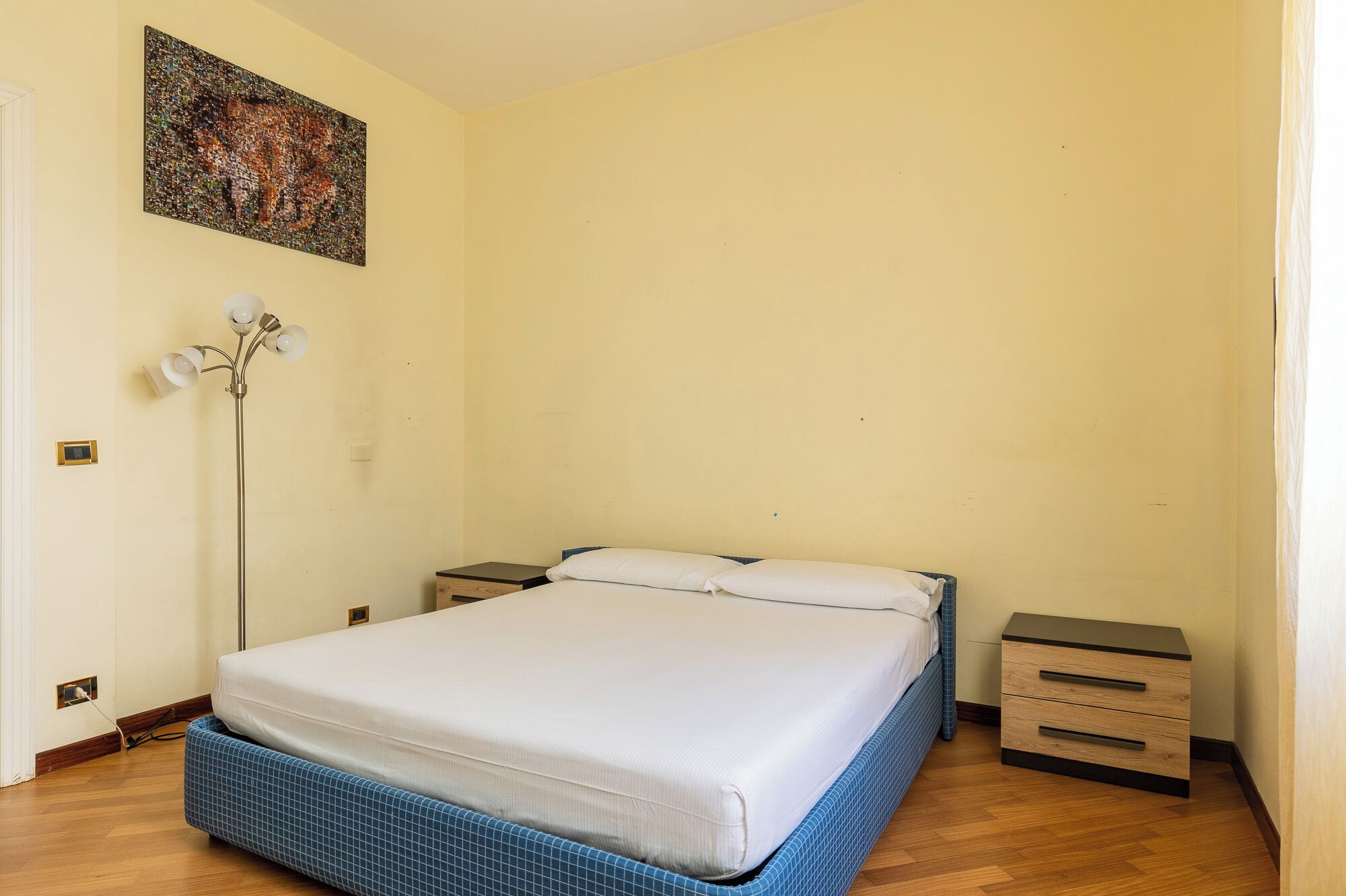 A bedroom with a bed, bedside tables, a floor lamp, and a painting on the wall.