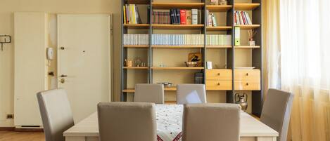 Ein Zimmer mit einer Holzregal voller Bücher, einem Schreibtisch mit Stuhl und einem Esstisch mit Stühlen.