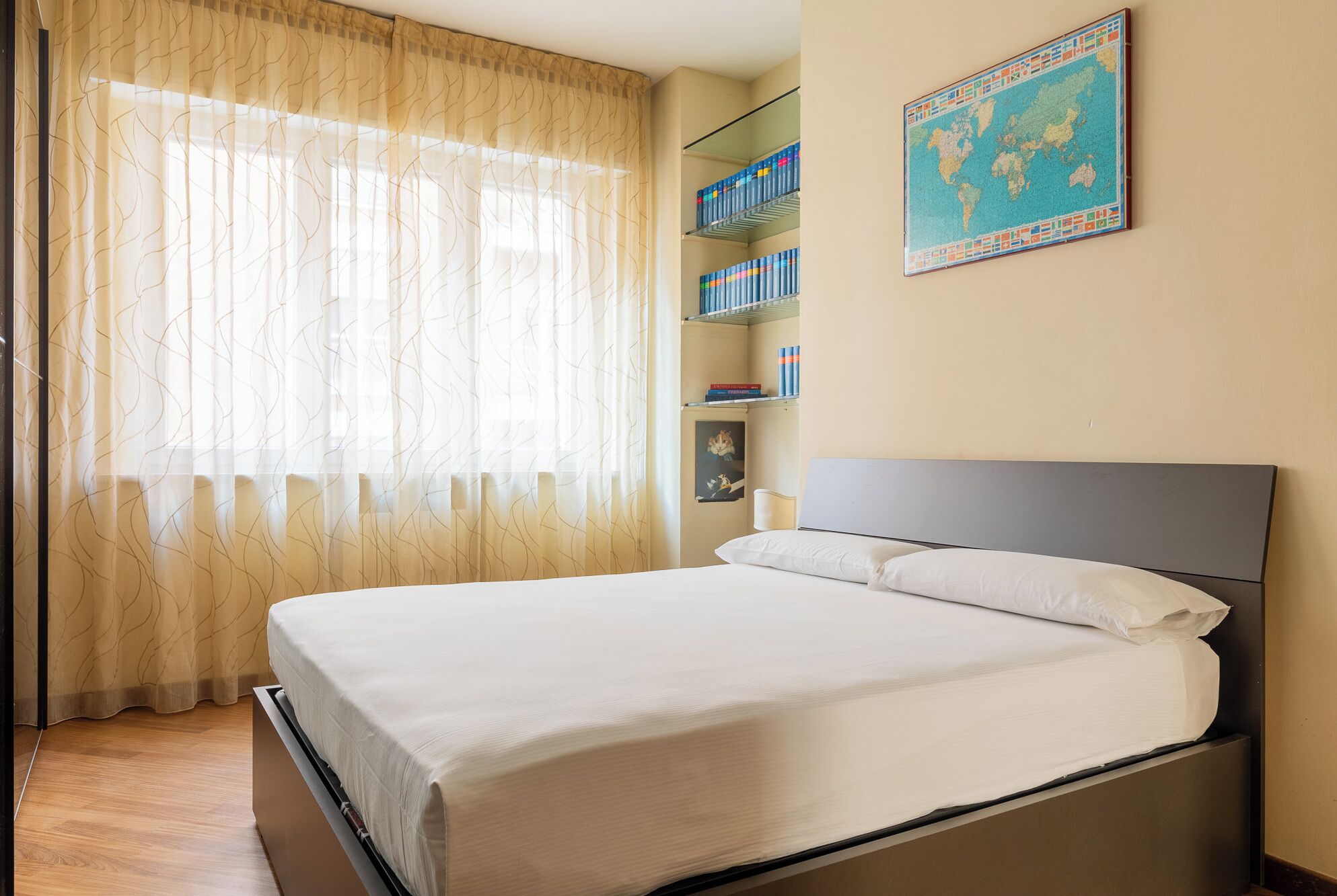 A neatly made bed in a hotel room with a large window, a bookshelf with books, and a world map on the wall.