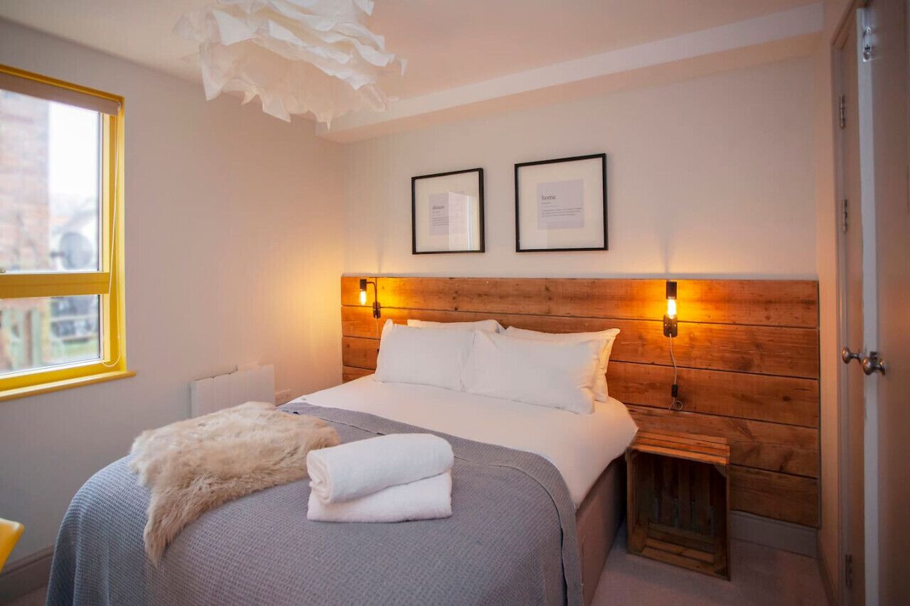 A neatly arranged bedroom with a wooden headboard, a bed with white linens, two framed certificates on the wall, and a small bedside table with a lamp.