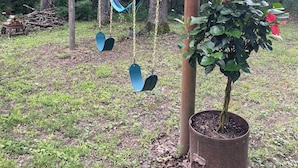 Un conjunto de columpios con hamaca en zona boscosa, con una planta en maceta cerca.