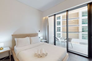 A hotel room with a bed, a nightstand, a lamp, a balcony with a view of the building, and a table with chairs.