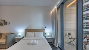 Une chambre d’hôtel moderne avec un grand lit, deux tables de chevet avec des lampes, un canapé et un balcon avec des chaises.
