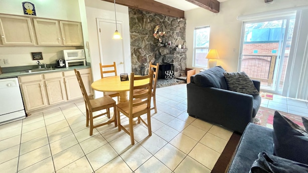 A living room with a sofa, a dining table with chairs, a kitchen area with cabinets and appliances, a stone fireplace, and a view through a glass door. - NEW LISTING! Panoramic Views At Panorama! (Breckenridge)