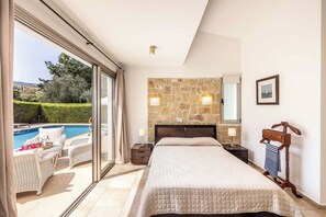 A bedroom with a large bed, a stone wall, a view of a swimming pool, and a wooden chair.