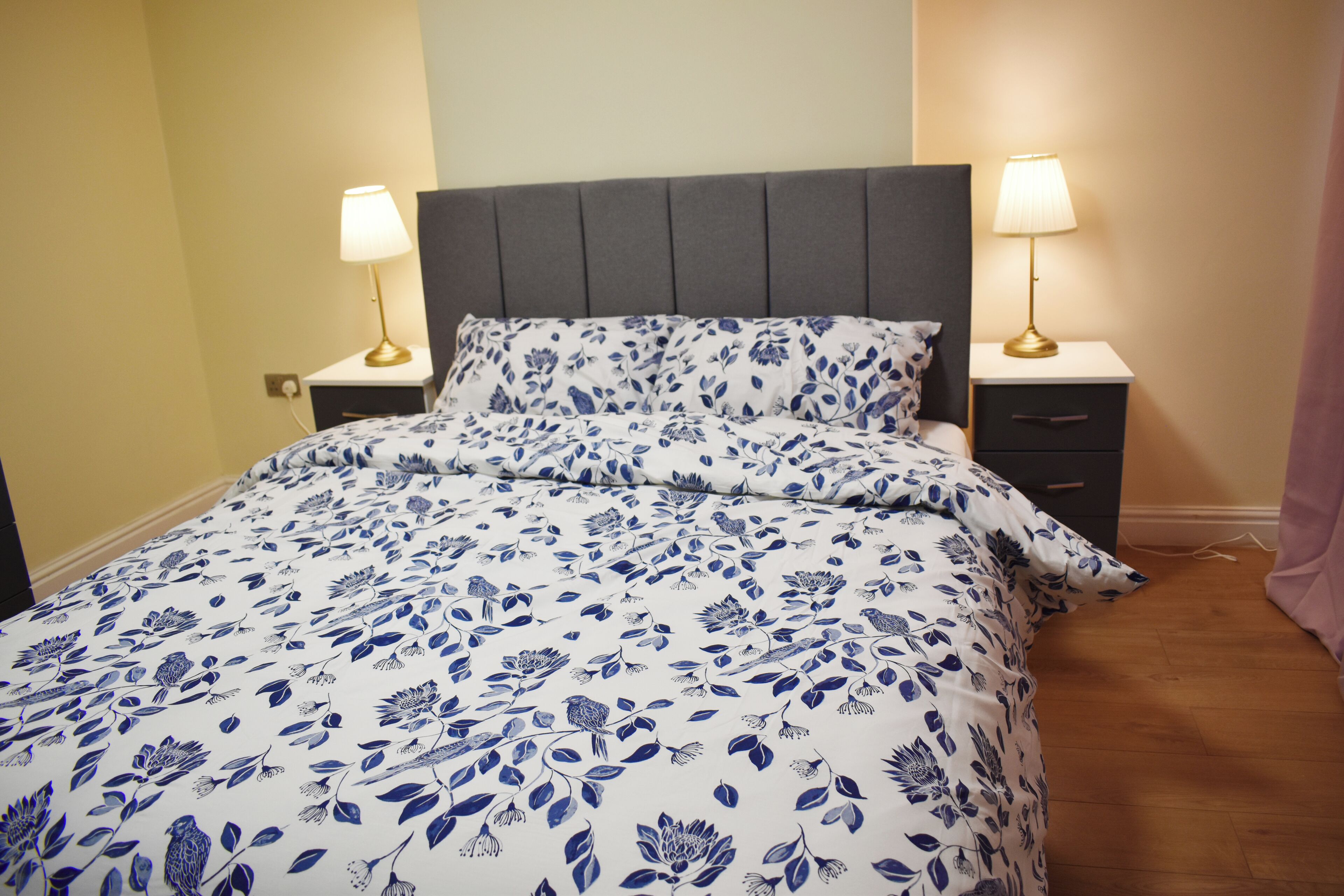 Un lit avec une couvre-lit à motifs floraux, deux tables de chevet avec des lampes et une tête de lit grise.