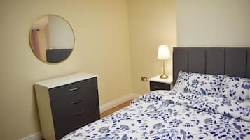 A bedroom with a bed, a bedside table with a lamp, a dresser, and a round mirror on the wall.