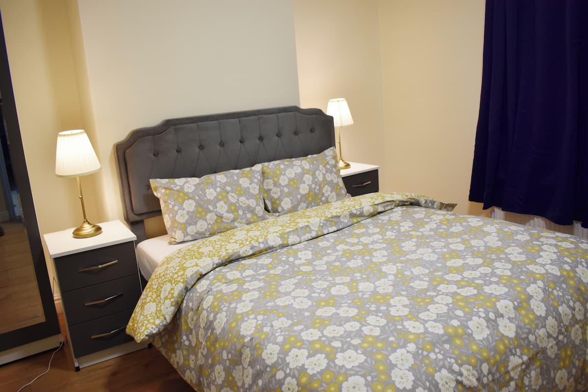 A bedroom with a grey upholstered headboard, two bedside tables with lamps, a floral patterned bedspread, and a dark blue curtain.