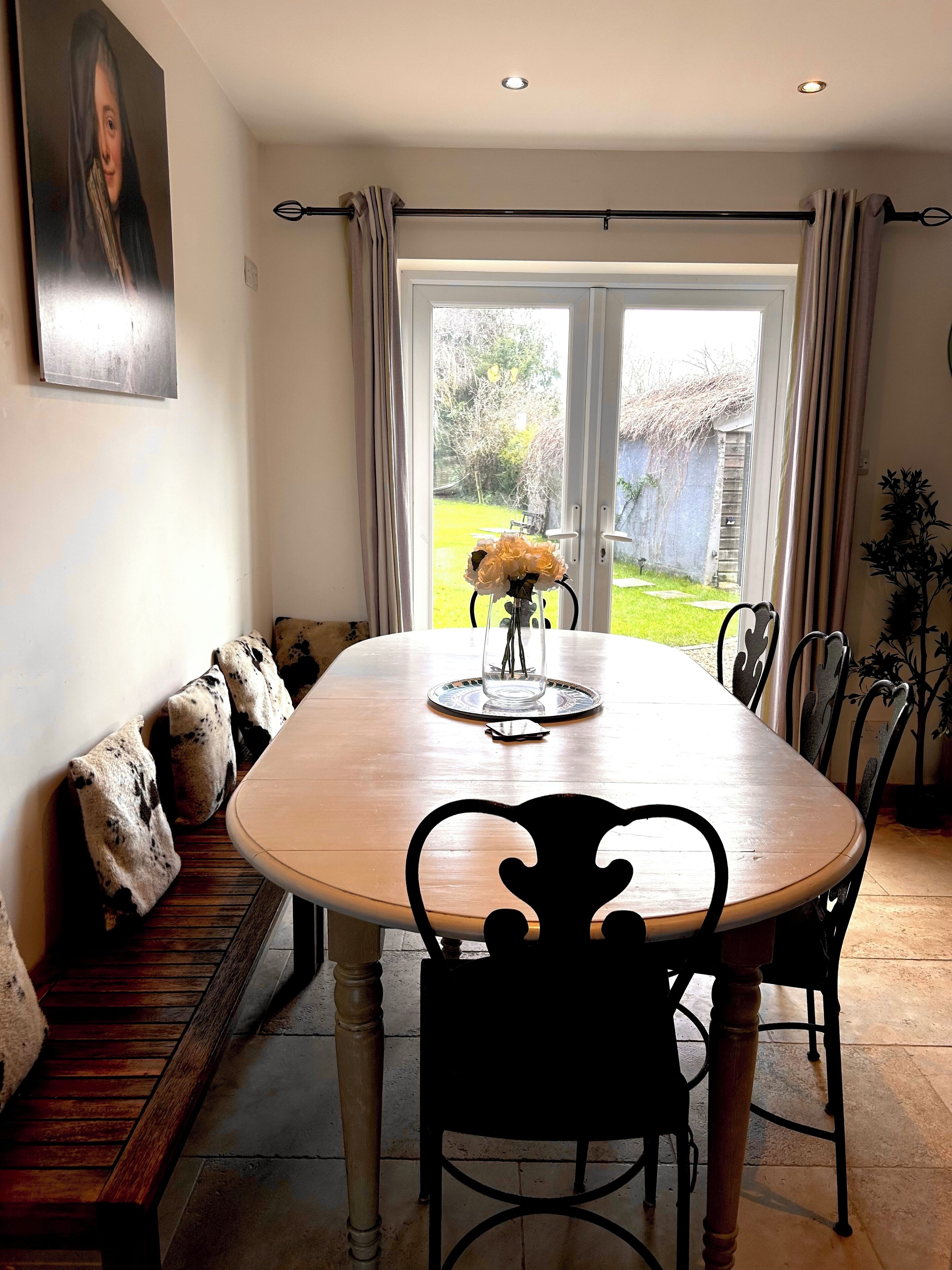 A dining area with a wooden table, black chairs, a bench with cushions, a vase with flowers, and a view of a garden through a glass door.