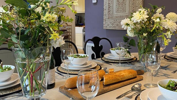Una mesa de comedor dispuesta con platos, vasos, cubiertos, pan y centros de mesa con flores.