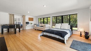 A spacious bedroom with a large bed, a foosball table, a desk, and a window with a view of greenery.