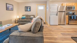 A modern living room with a grey sofa, a wooden coffee table, and a blue rug. There is a kitchen area with stainless steel appliances and wooden cabinets. - Wilderness Escape 2 bedroom 1 bath 4 guests (Sterling)