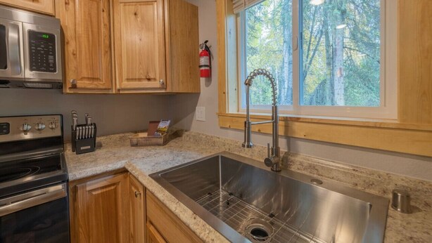A kitchen with wooden cabinets, a stainless steel sink, a microwave, and a window with a view of trees. - Wilderness Escape 2 bedroom 1 bath 4 guests (Sterling)