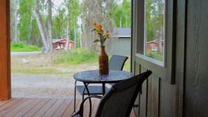 A wooden deck with a round table and chairs, a vase with flowers, and a view of a red building and trees.