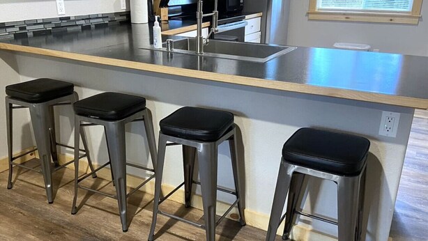 A kitchen with a stainless steel countertop, four bar stools, and a window with blinds.