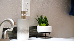 A bathroom with a modern faucet, a white sink, a potted plant, and a wall-mounted electrical outlet.