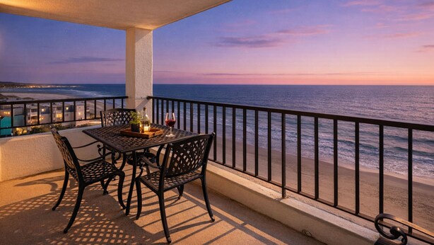 Een balkon met een tafel en stoelen, met uitzicht op een strand en zee bij zonsondergang.