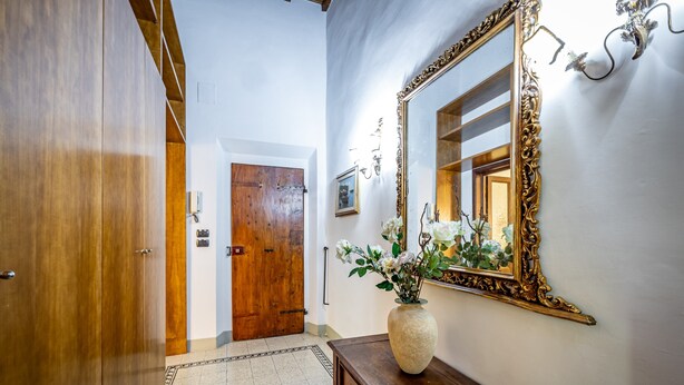 Un pasillo con puertas de madera, un espejo grande y ornamentado, y un jarrón con flores sobre una consola de madera.