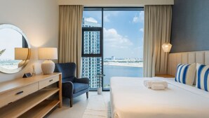 A hotel room with a large bed, a desk with a mirror, a chair, and a view of a cityscape and water.