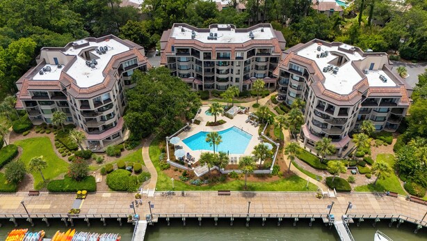 A modern apartment complex with a central swimming pool, surrounded by lush greenery and a dock with boats. - Sunset Rentals | Yacht Club 7548 | Shelter Cove (Hilton Head Island)