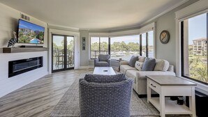 A modern living room with a flat-screen TV, a fireplace, a sofa, a coffee table, and large windows offering a view of the city. - Sunset Rentals | Yacht Club 7548 | Shelter Cove (Hilton Head Island)
