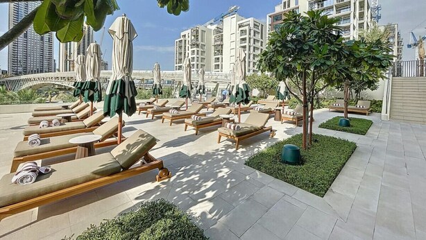 A rooftop lounge area with sun loungers and umbrellas, surrounded by greenery and modern buildings in the background.