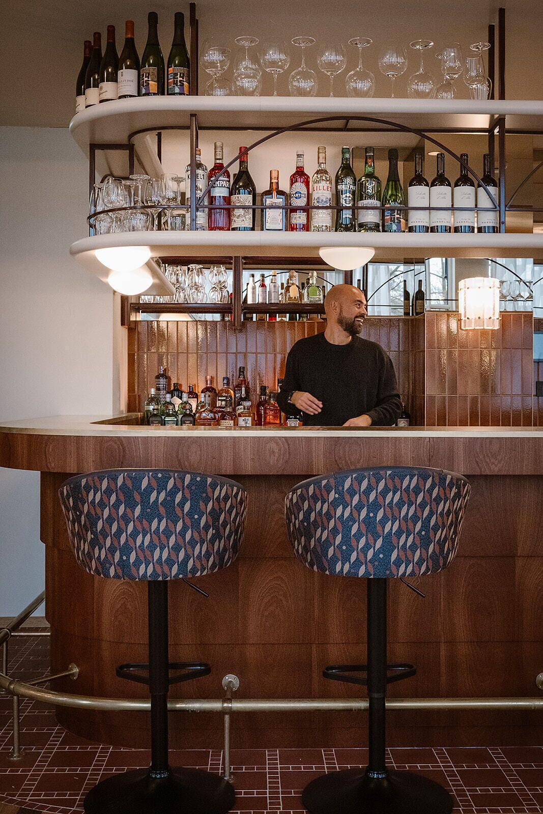 Een bar met twee gestreepte krukken, planken met flessen en glazen, en een bartender.