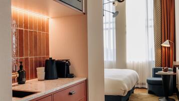 Habitación de hotel con baño que cuenta con un muro de azulejos, un lavamanos y artículos de tocador. La habitación tiene una cama, una silla y una mesita con lámpara.