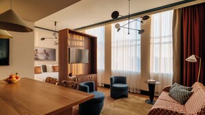 A modern hotel room with a wooden dining table, a sofa, a blue armchair, and a TV mounted on the wall. - De Plesman Hotel The Hague (The Hague)