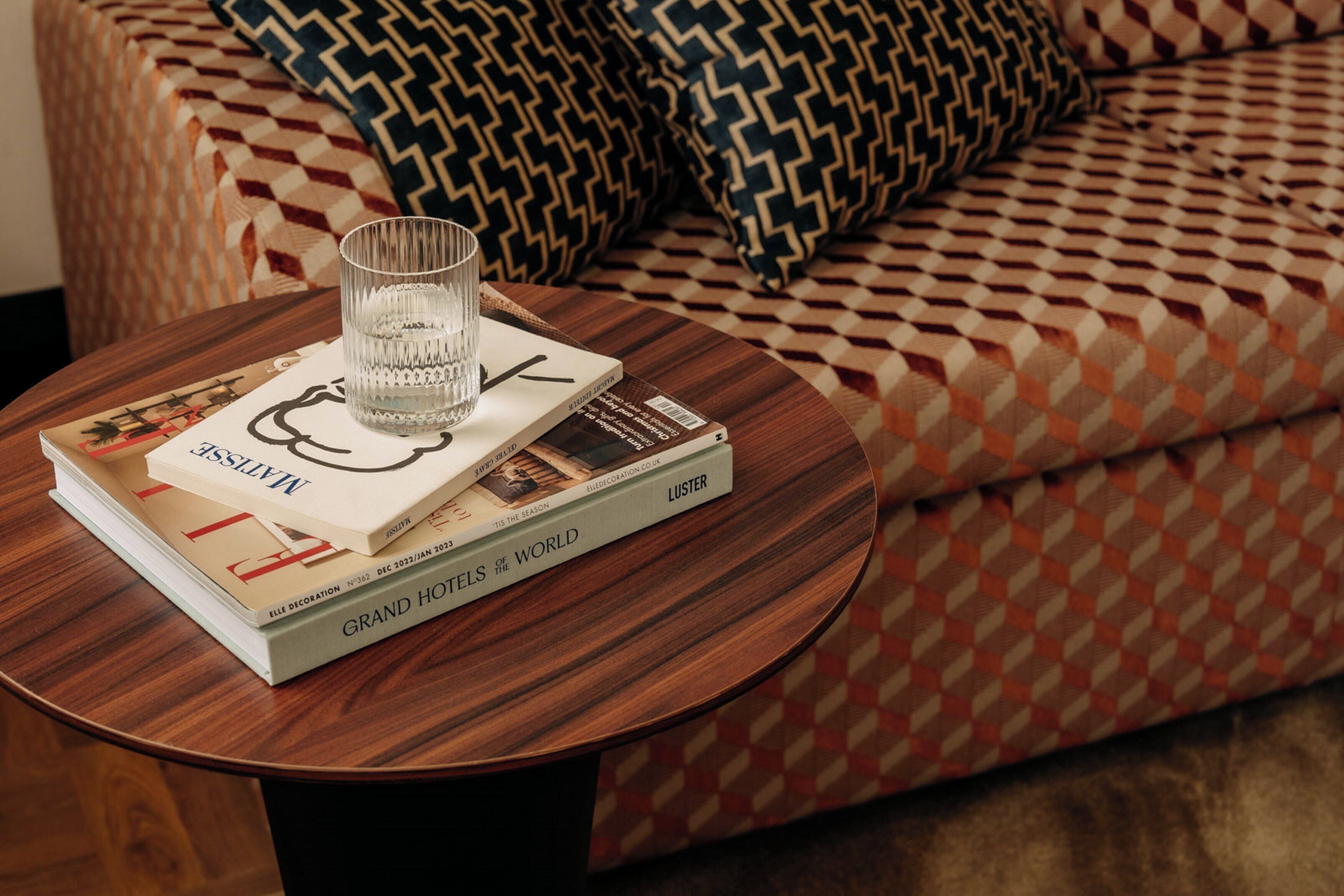 A round wooden table with a glass of water, a book titled 'Grand Hotels of the World', and a patterned cushion in the background.