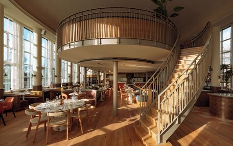 A spacious restaurant with a central circular seating area, wooden flooring, and a staircase with a railing.