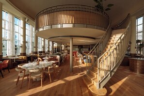 A spacious restaurant with a central circular seating area, wooden flooring, and a staircase with a railing. - De Plesman Hotel The Hague (The Hague)