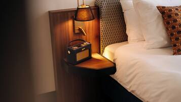 Una cama bien hecha con ropa blanca y almohadas estampadas, una lámpara de noche y una radio pequeña sobre una mesita de madera.