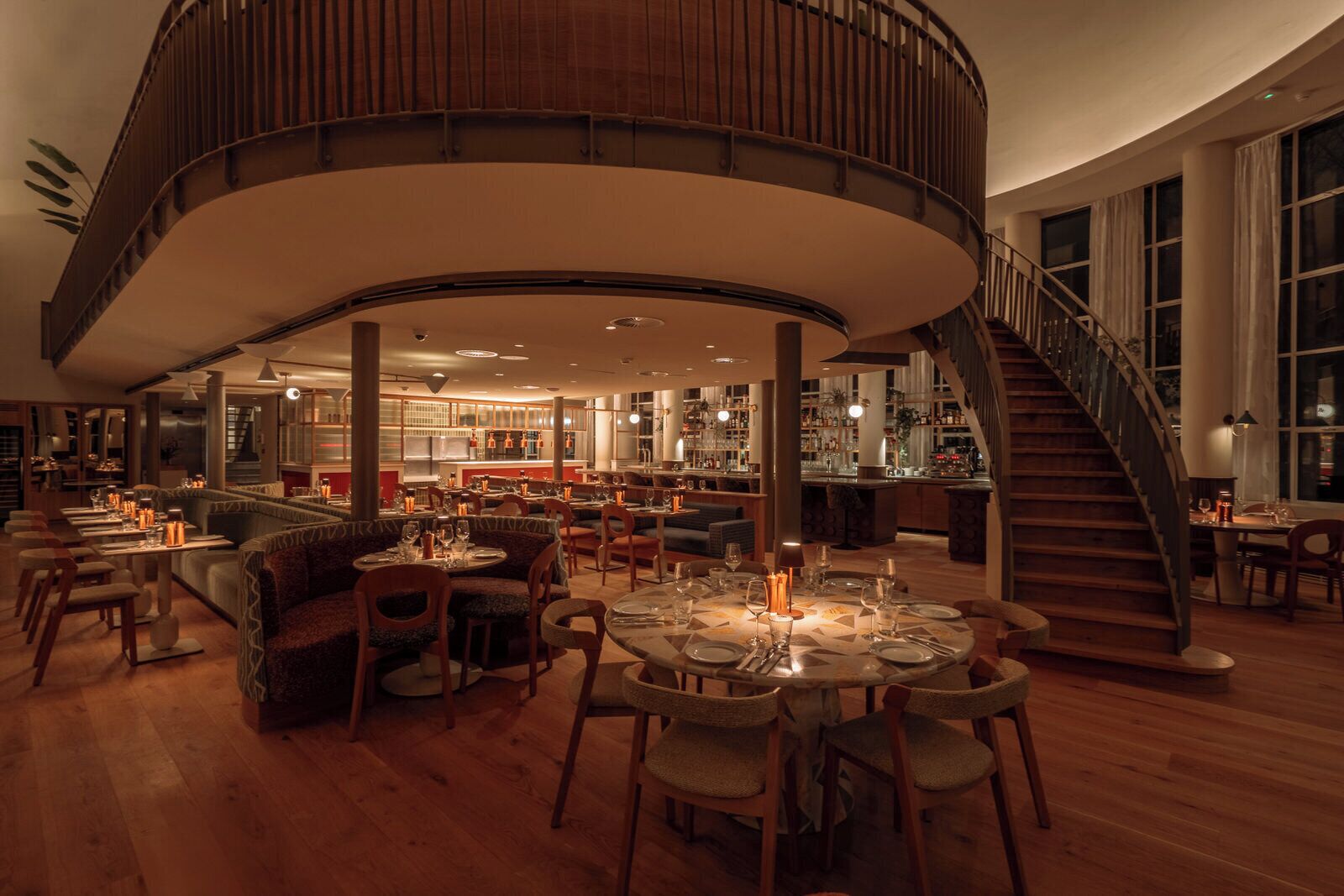 A spacious, elegantly designed restaurant with a central seating area, a bar, and a spiral staircase.