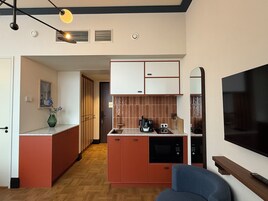 Une cuisine compacte équipée d’un four à micro-ondes, d’une cuisinière et d’un évier. Un téléviseur est fixé au mur et il y a un petit coin salon avec une chaise bleue.
