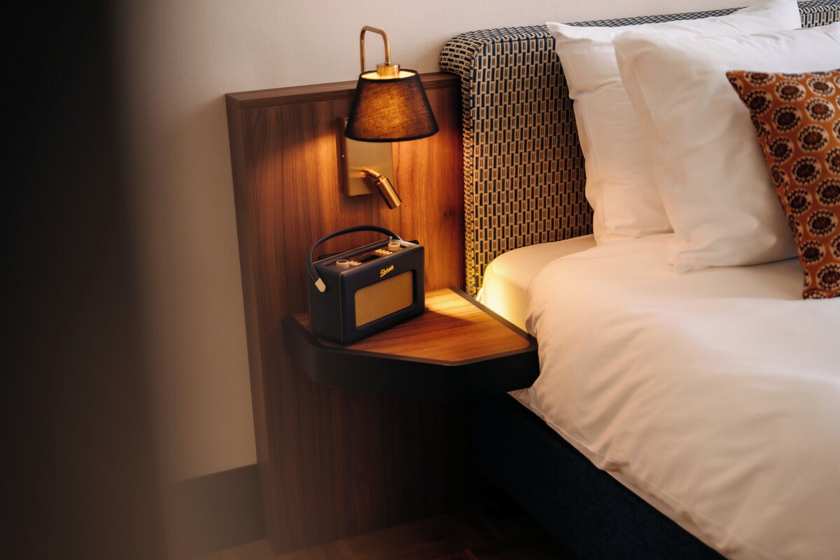 A neatly made bed with white linens and patterned pillows, a bedside lamp, and a small radio on a wooden bedside table.