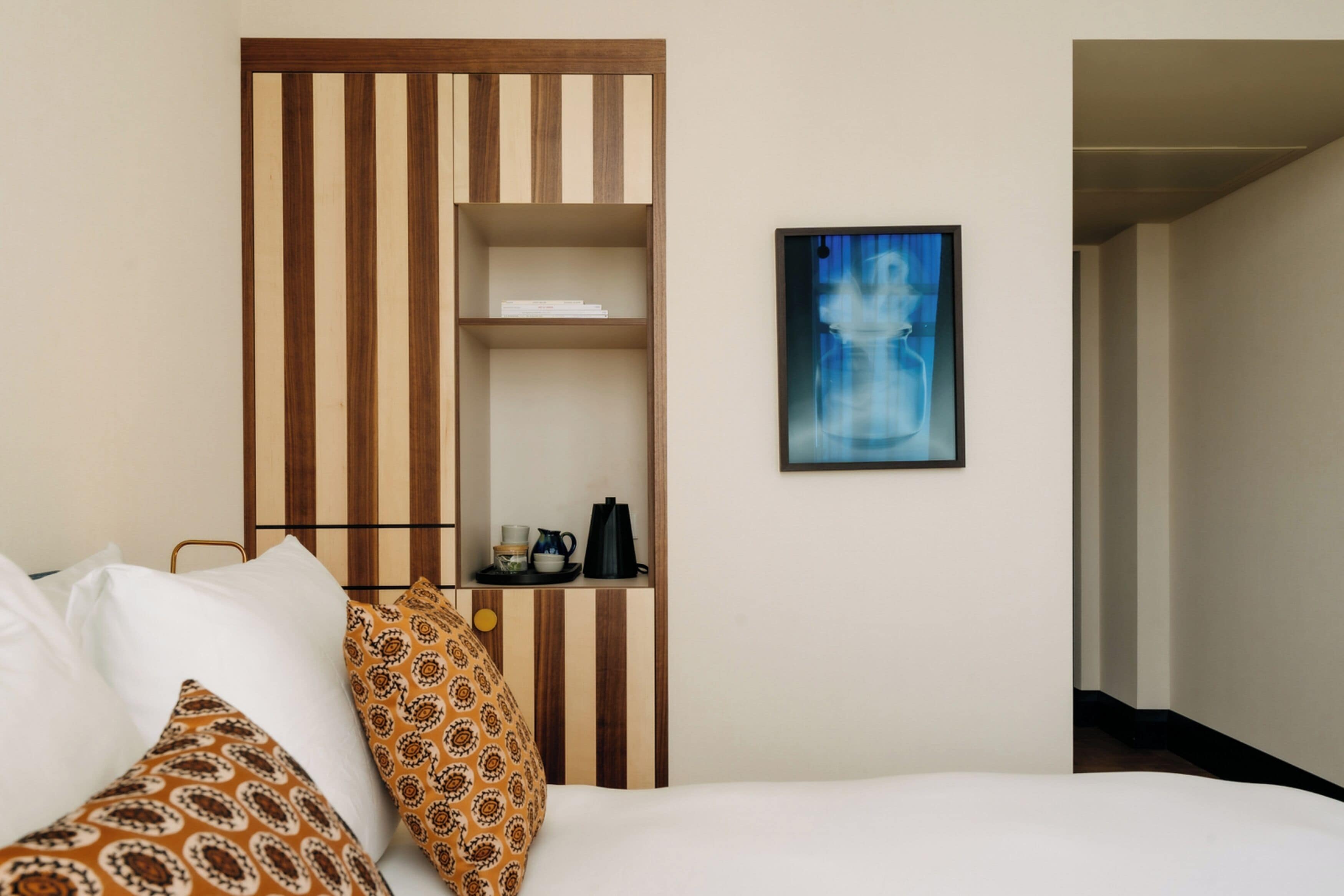 Camera d'albergo con un letto, una scaffalatura in legno, un quadro astratto incorniciato e vista su un corridoio.