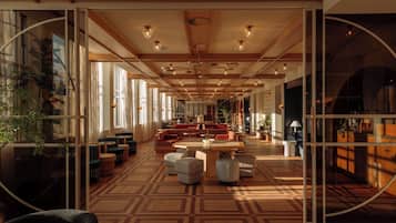 A spacious hotel lobby with a high ceiling, wooden beams, and a patterned floor. There are multiple seating areas with tables and chairs, and a large window on one side.