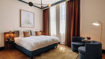 Una habitación de hotel moderna con una cama grande, una zona de estar con un sillón, un escritorio con lámpara y un amplio ventanal con cortinas.