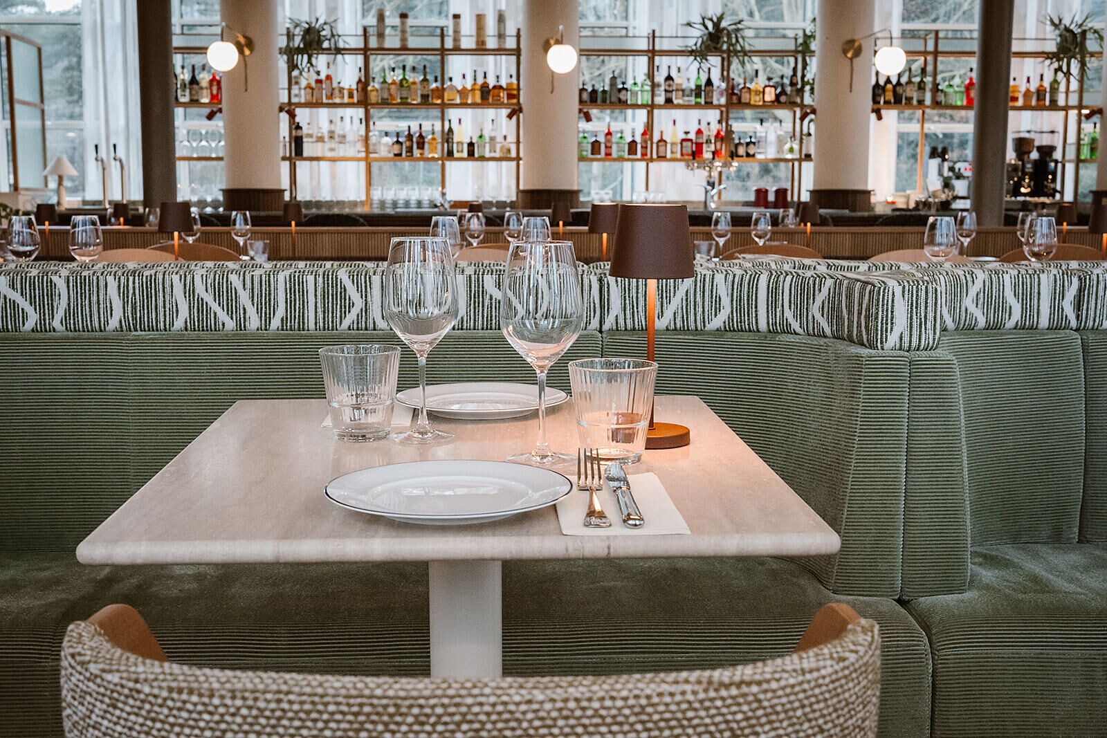Un ambiente de restaurante con una mesa dispuesta para dos, un bar al fondo y un banco tapizado verde con diseño.