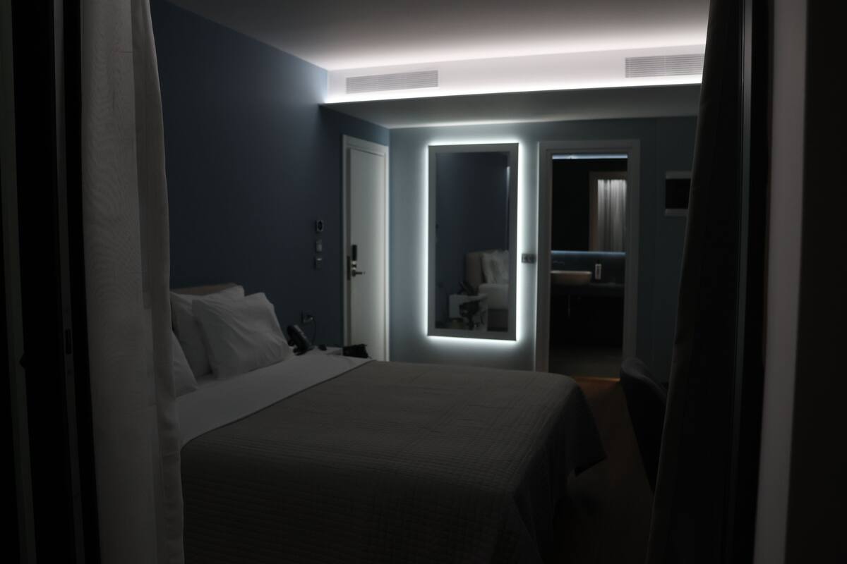 Habitación de hotel con cama, mesita de noche y espejo. Hay una puerta que da a un baño.