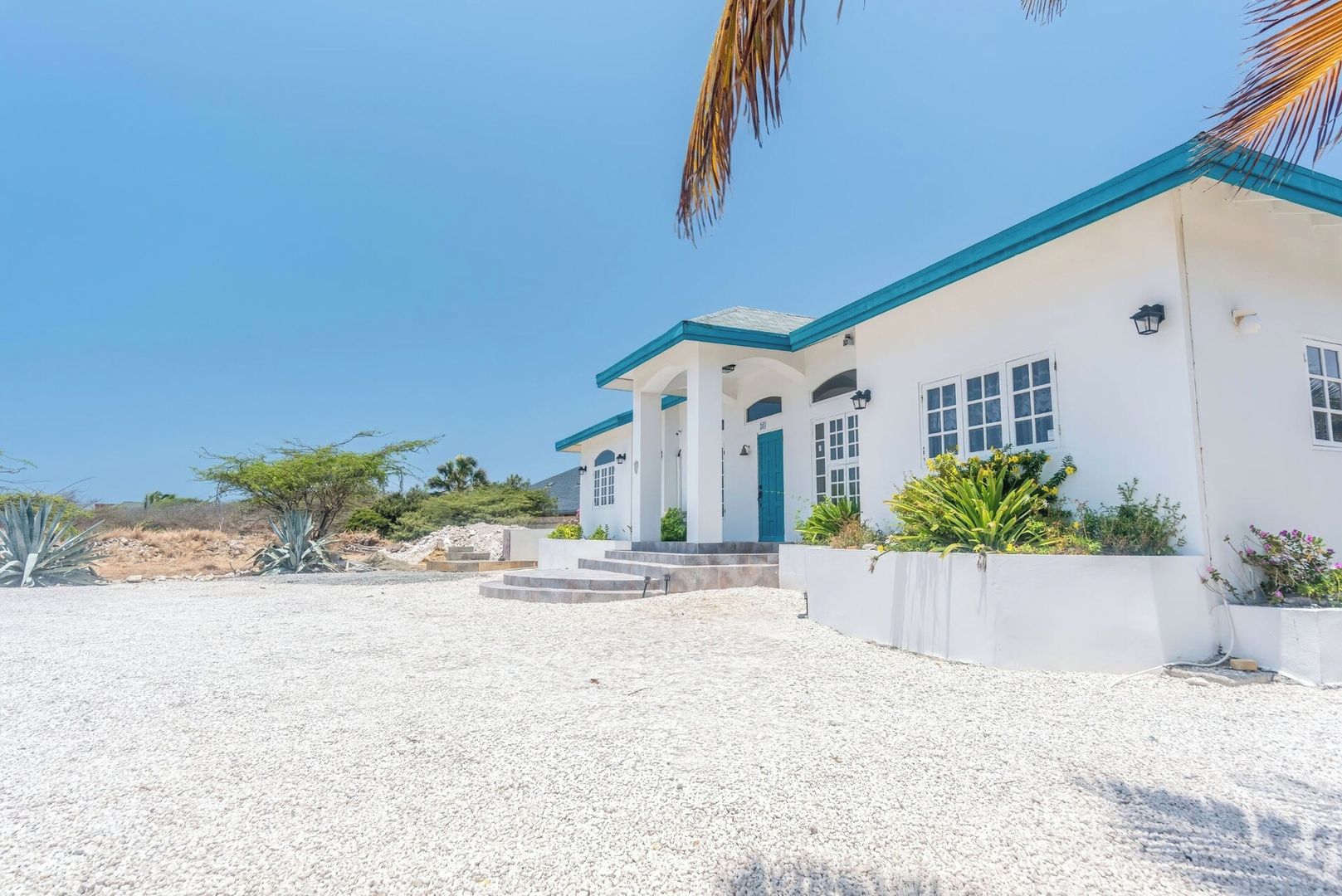 A white building with blue trim and a covered porch, surrounded by gravel and sparse vegetation.