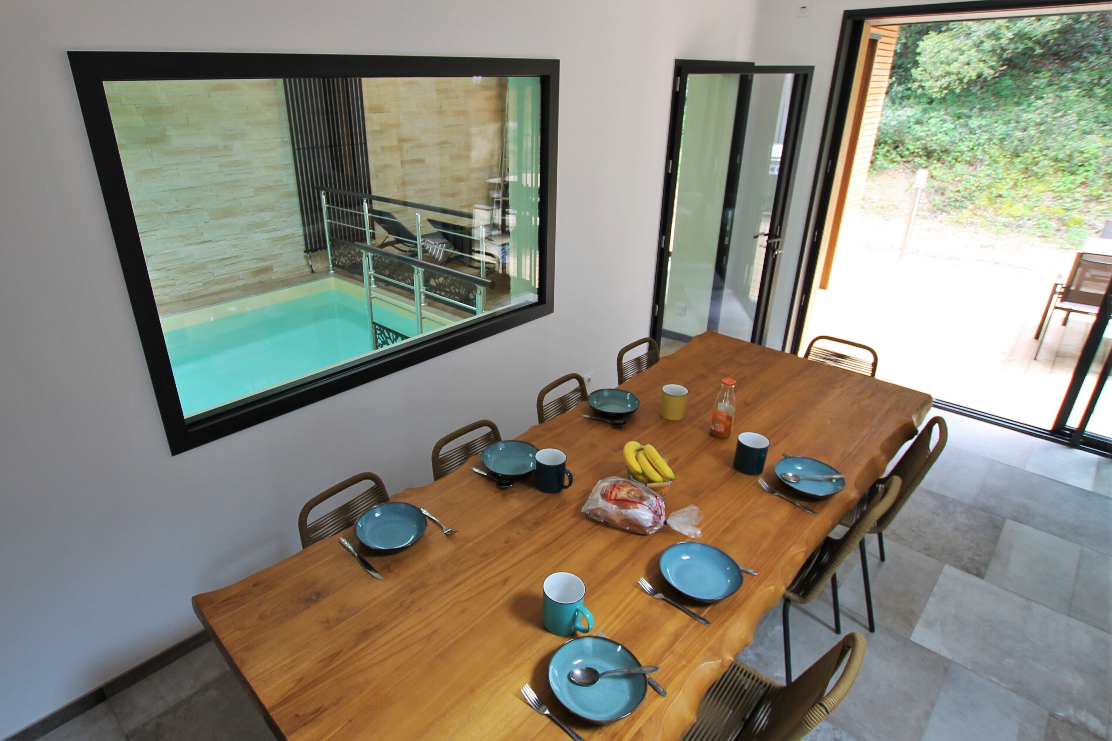 Un comedor con una mesa de madera dispuesta para seis personas, rodeada de sillas. Un amplio ventanal muestra una piscina y zona de estar al aire libre.