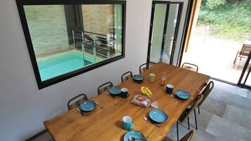 Un comedor con una mesa de madera dispuesta para seis personas, rodeada de sillas. Un amplio ventanal muestra una piscina y zona de estar al aire libre.