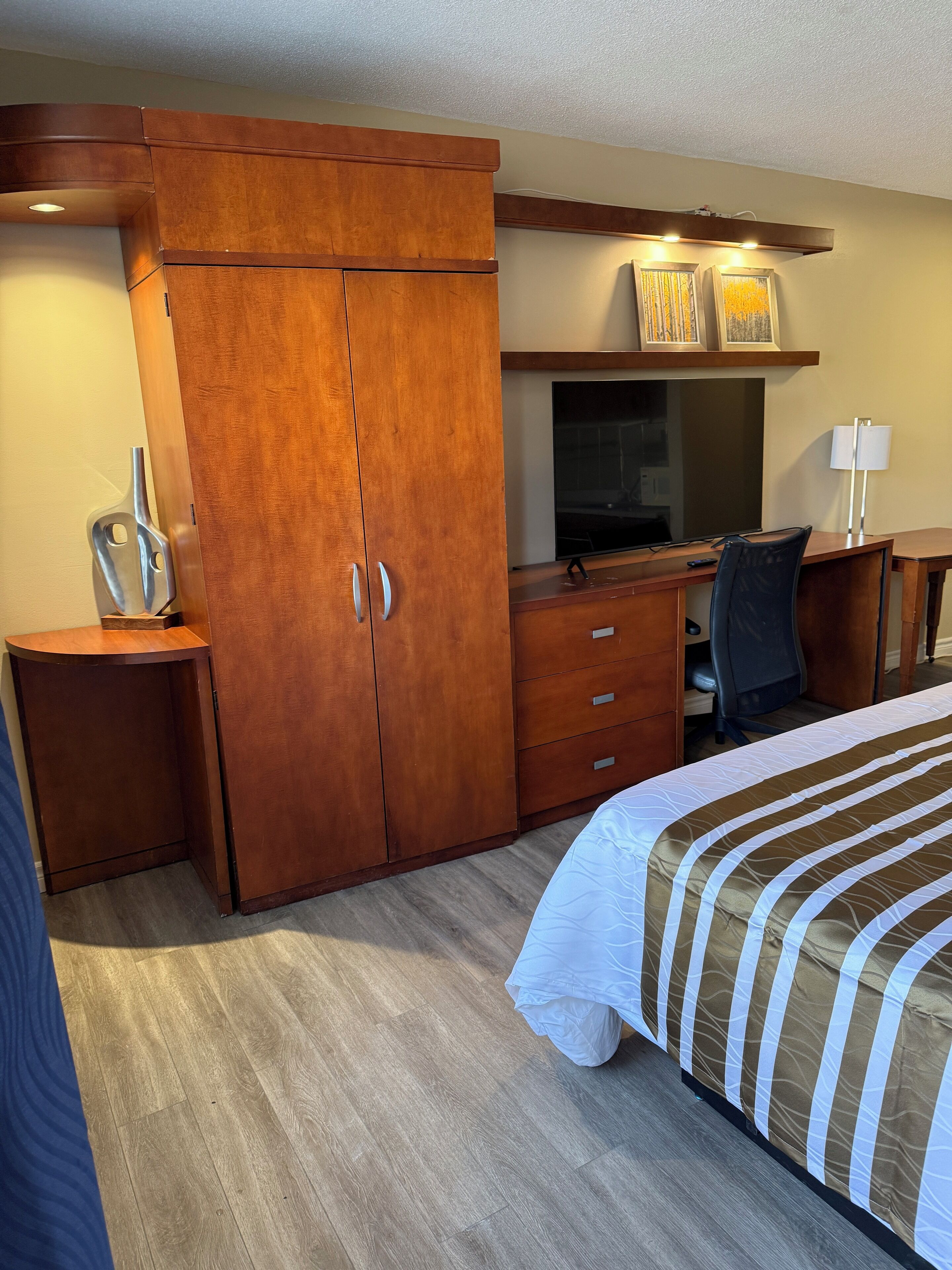 Ein Hotelzimmer mit Holzschrank, Schreibtisch mit Computer, Fernseher und Bett mit gestreifter Bettwäsche.