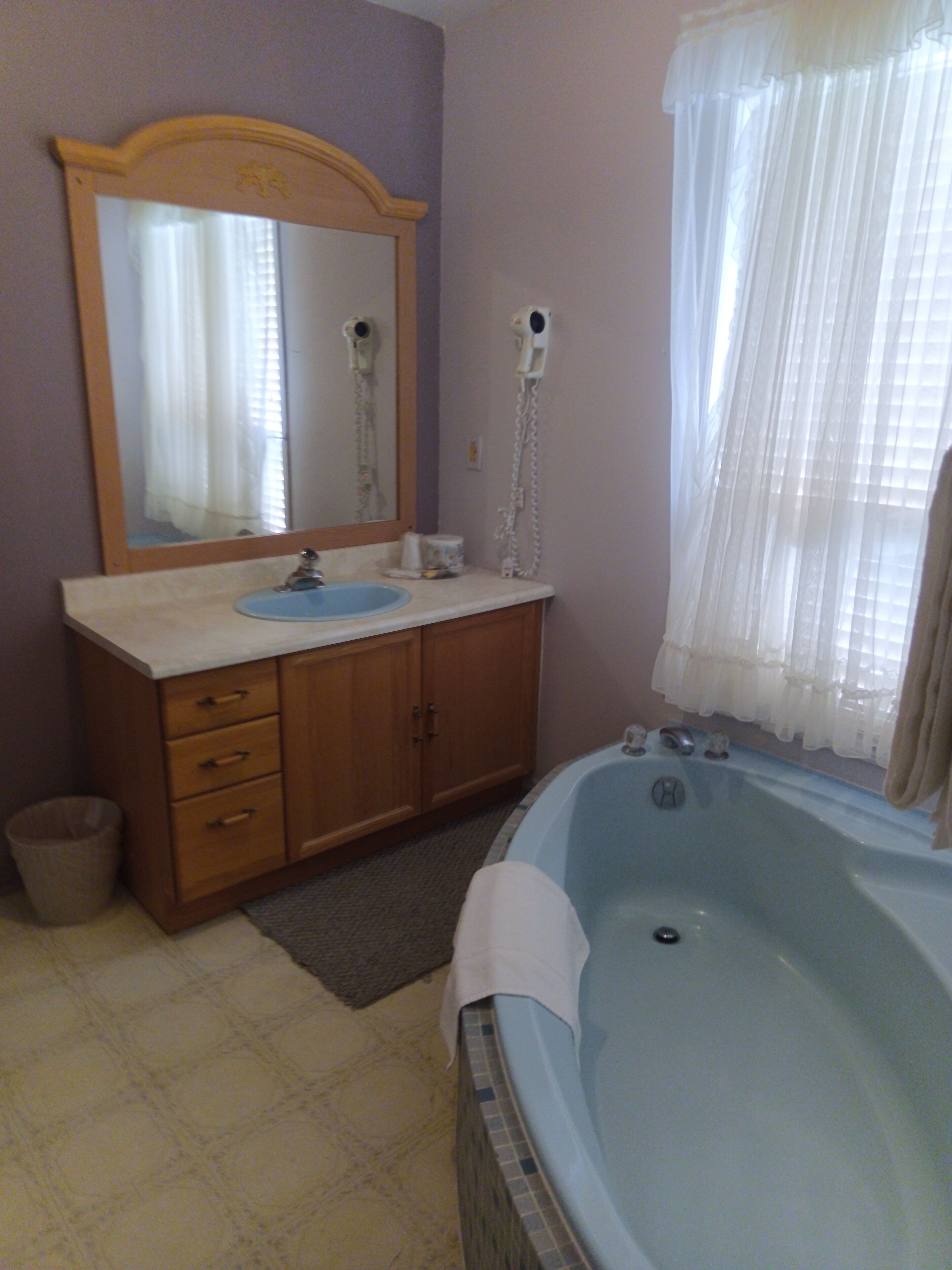 Une salle de bain avec un meuble-lavabo en bois, un lavabo bleu, une grande baignoire et un miroir sur lequel est fixé un sèche-cheveux au mur.
