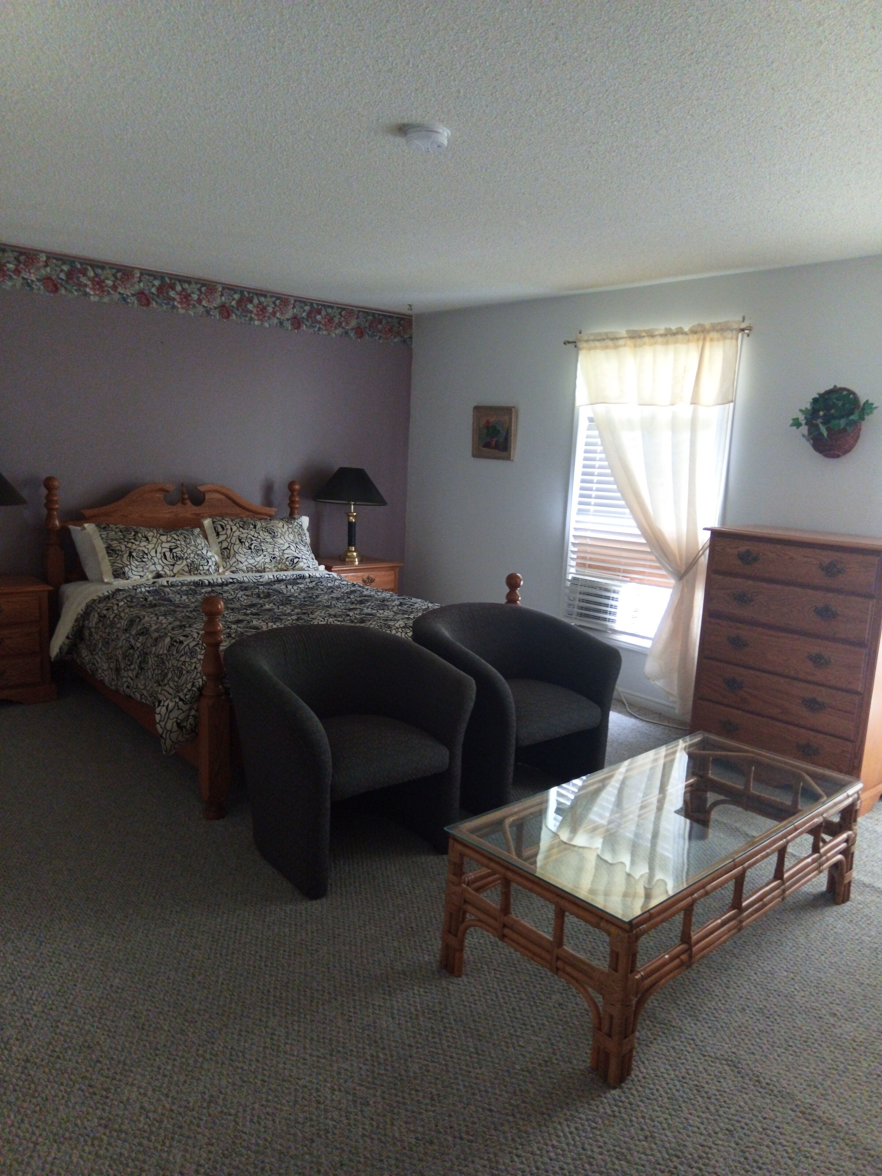 A bedroom with a bed, two armchairs, a dresser, a nightstand, a lamp, a window with curtains, and a ceiling light.