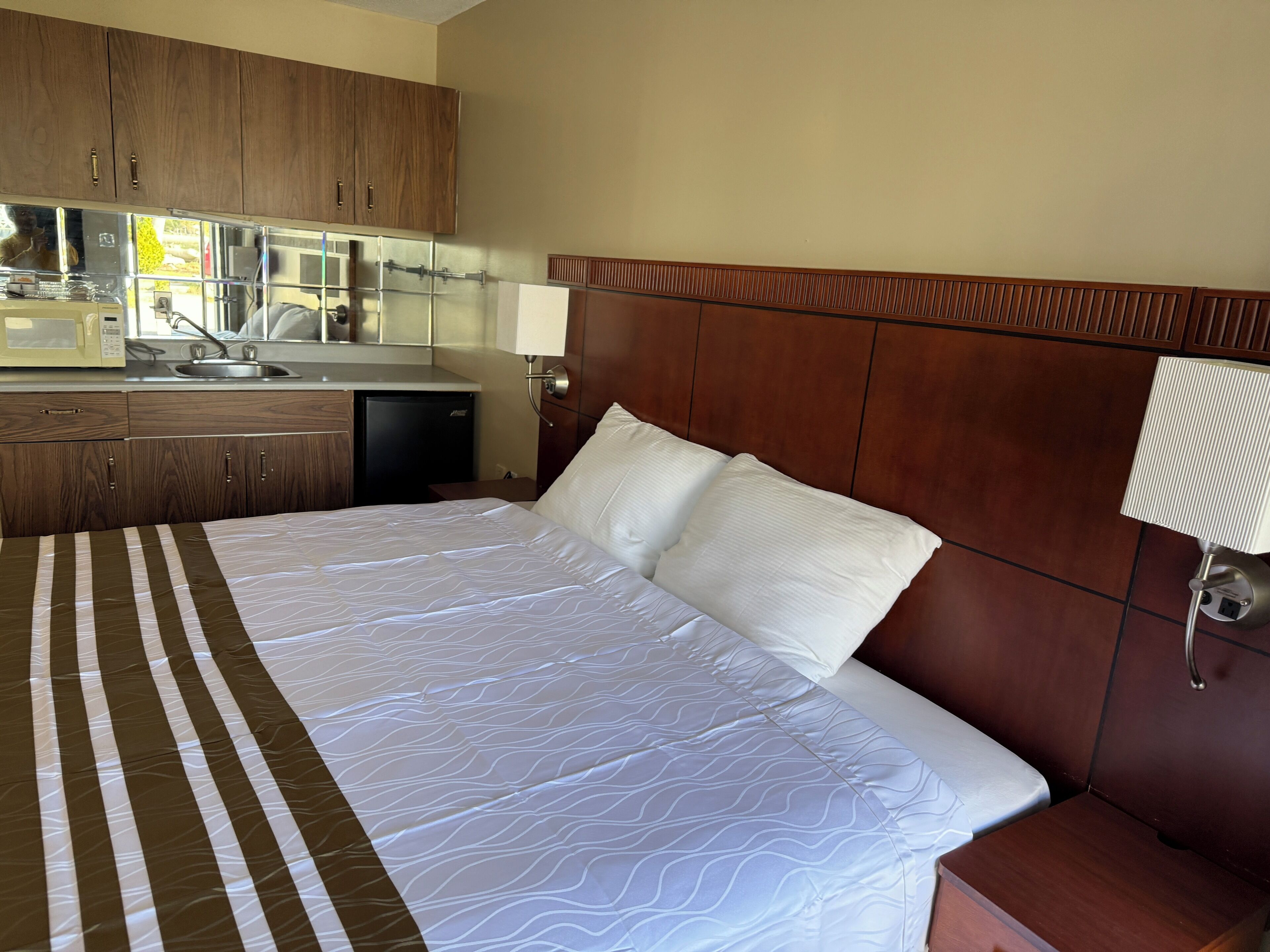A hotel room with a bed, a wooden headboard, a small kitchenette with a microwave, sink, and refrigerator.