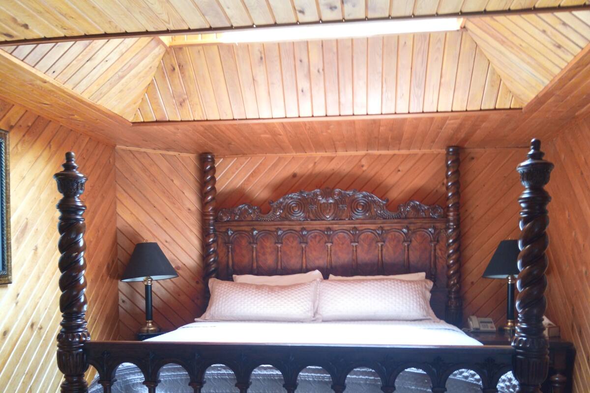 A bedroom with a wooden ceiling, a four-poster bed, two bedside lamps, and a glass-topped table.